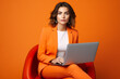 Beautiful business woman wearing orange business suit with a laptop at armchair, isolated on orange background