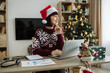 © sofiko14 - Caucasian businesswoman wearing Santa hat sitting at home near xmas tree and working on laptop during the holiday season, typing on computer keyboard.