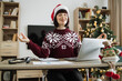 © sofiko14 - Front view of female doing yoga exercise while sitting at desk with gadgets and papers at decorated with Christmas tree home. Caucasian woman in Santa hat relaxing with closed eyes after work at home.