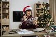 © sofiko14 - Smiling happy adult in Santa hat showing okay gesture while holding smartphone in living room decorated with Christmas tree. Businesswoman greeting video call participants during working time.