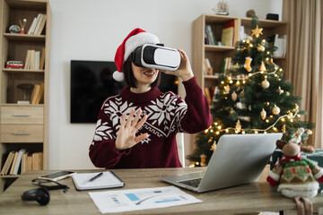 Caucasian young adult businesswoman wearing Santa hat using VR glasses touching air and gesturing, sitting at table with gadgets at home near xmas tree during the holiday season.