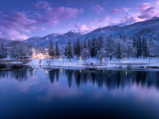 Wall Mural - aerial view of lake, snowy alpine mountains, pine trees in snow, reflection in water, purple sky wit