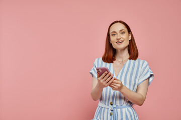 Wall Mural - cheerful stylish woman in striped dress holding smartphone and looking at camera on pink backdrop