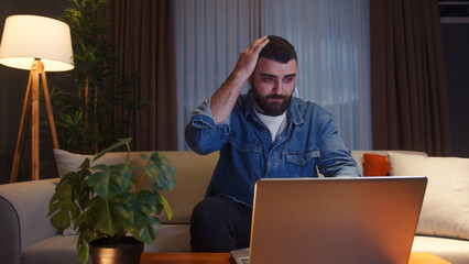 Wall Mural - Young adult man sitting on sofa at home while using laptop having anxiety and stress. Upset young man reacting to loss, bad news on laptop