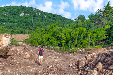 Male traveler walks through a forest plot cleared for construction in the mountains near Budva, Montenegro. Young hiker with a backpack among boulders and cobblestones on the backdrop of a green wood