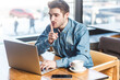 © khosrork - Portrait of attractive bearded young man freelancer in blue jeans shirt working on laptop, having video call, showing silent gesture. Indoor shot near big window, cafe background.