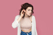 Portrait of serious curious woman with curly hair wearing casual style outfit keeps hand near ear, listening to something private, gossip. Indoor studio shot isolated on pink background.