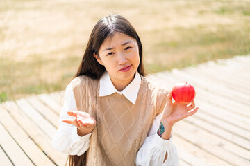 Wall Mural - Young Chinese woman with an apple at outdoors making doubts gesture while lifting the shoulders