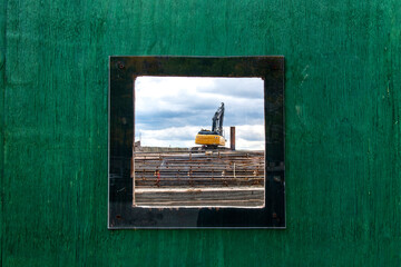 Viewing panel, porthole, or window in plywood wall around construction site in New York City, NY, USA with look on pile of rebar of wire mesh and bright yellow heavy duty crane on steel tracks