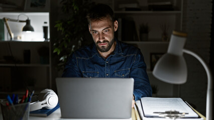 Driven young hispanic man, working long nights at office; focused, serious yet relaxed, this bearded professional navigates business on his laptop.