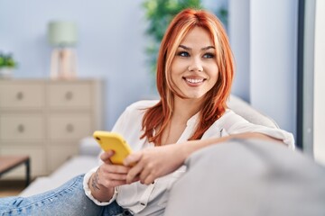 Wall Mural - Young caucasian woman using smartphone sitting on sofa at home
