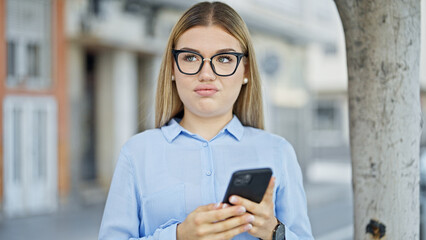 Wall Mural - Young blonde woman business worker using smartphone with serious expression at street