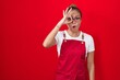 Young hispanic woman wearing waitress apron over red background doing ok gesture shocked with surprised face, eye looking through fingers. unbelieving expression.