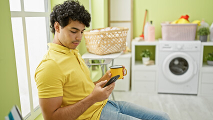 Sticker - Young latin man using smartphone drinking coffee waiting for washing machine at laundry room