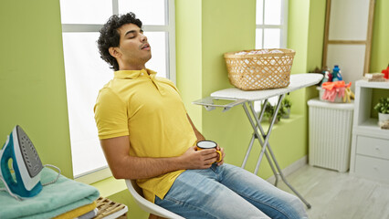 Sticker - Young latin man waiting for washing machine holding coffee sleeping at laundry room