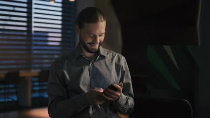 Carefree positive browsing phone mobile app Caucasian businessman at evening office using smartphone smiling happy business man entrepreneur scrolling cellphone check email work break message network