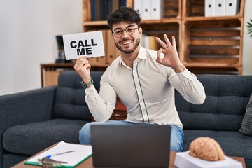 Canvas Print - Hispanic man with beard working at therapy office holding call me sign doing ok sign with fingers, smiling friendly gesturing excellent symbol