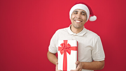 Poster - Young hispanic man smiling confident holding christmas present over isolated red background