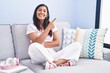 © Krakenimages.com - Young hispanic woman sitting on the sofa at home with a big smile on face, pointing with hand and finger to the side looking at the camera.