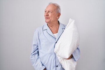 Poster - Senior man with grey hair wearing pijama hugging pillow looking away to side with smile on face, natural expression. laughing confident.