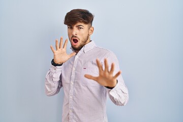 Canvas Print - Arab man with beard standing over blue background afraid and terrified with fear expression stop gesture with hands, shouting in shock. panic concept.