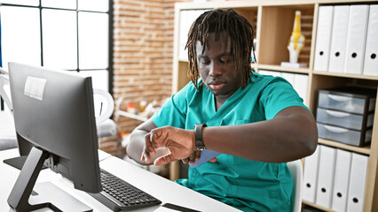 Wall Mural - African american man doctor using computer looking watch with serious face at the clinic