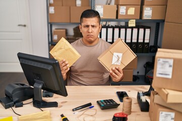 Poster - Hispanic young man working at small business ecommerce holding packages skeptic and nervous, frowning upset because of problem. negative person.