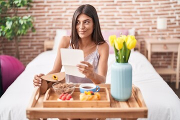 Canvas Print - Brunette young woman eating breakfast in the bed reading a letter smiling looking to the side and staring away thinking.