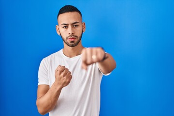 Sticker - Young hispanic man standing over blue background punching fist to fight, aggressive and angry attack, threat and violence