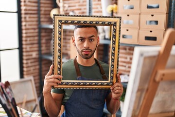 Poster - Young hispanic man sitting at art studio with empty frame skeptic and nervous, frowning upset because of problem. negative person.