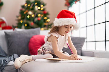 Sticker - Adorable caucasian girl drawing on notebook sitting on sofa by christmas tree at home