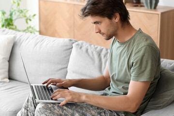 Wall Mural - Young male soldier with laptop sitting on sofa at home