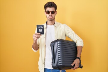 Wall Mural - Young hispanic man holding suitcase going on summer vacation holding italian passport depressed and worry for distress, crying angry and afraid. sad expression.