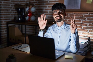 Canvas Print - Young hispanic man with beard working at the office at night showing and pointing up with fingers number seven while smiling confident and happy.