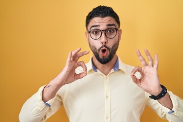 Poster - Hispanic young man wearing business clothes and glasses looking surprised and shocked doing ok approval symbol with fingers. crazy expression