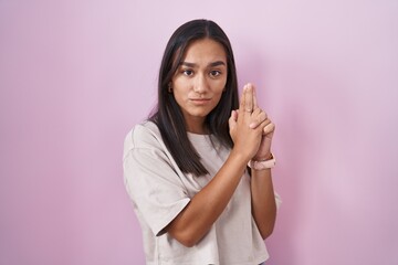 Wall Mural - Young hispanic woman standing over pink background holding symbolic gun with hand gesture, playing killing shooting weapons, angry face