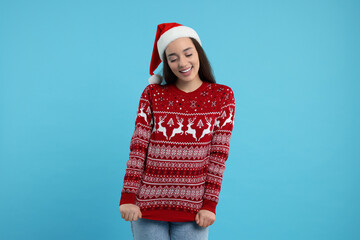 Poster - Happy young woman in Santa hat showing Christmas sweater on light blue background