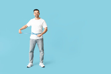 Wall Mural - Handsome sporty happy man measuring his waist on blue background. Weight loss concept