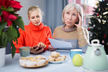 Wall Mural - Mature woman and her adult daughter, who came to visit her before Christmas, study an important document sitting at a table, ..dialing a number on a mobile phone that is listed there