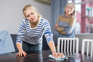 Elderly mother is unhappy with the way her adult daughter dusts the table in the room