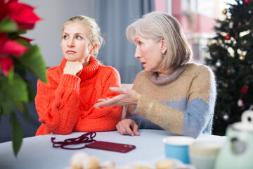 Wall Mural - Mature mother explains something to her adult thoughtful daughter, while sitting at the table in the room