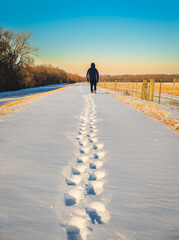 View of man walking away from camera in open field and leaving footprints in fresh snow at sunset in winter in Midwest