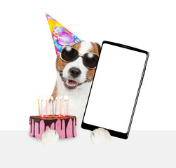 Wall Mural - Jack russell terrier puppy wearing sunglasses and party cap holds cake with lots burning candles and big smartphone with blank screen in it paw above empty white banner. isolated on white background