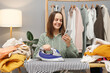 © sementsova321 - Cheerful brown haired woman wearing knitted shirt ironing clothing while sitting in her wardrobe using mobile phone scrolling online while doing household chores