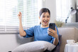 © David - Young Happy Asian woman smiling holding Phone with fist hand and excited for success on sofa at home.