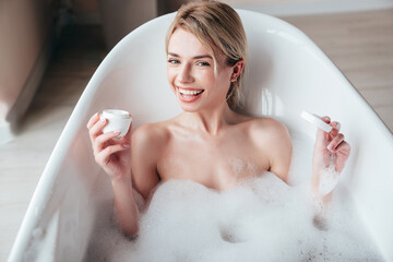 Wall Mural - Portrait of young beautiful sexy woman having fun while lying in bathtub full of foam at home. Charming smiling model relaxing in luxury bath interior. Female enjoying beauty and skincare day