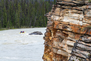 View over Athabasca River flowing from nearby Athabasca Falls, Jasper National Park, AB, Canada, with white-water raft in distance on river and river shore with colorful rock formations of limestone