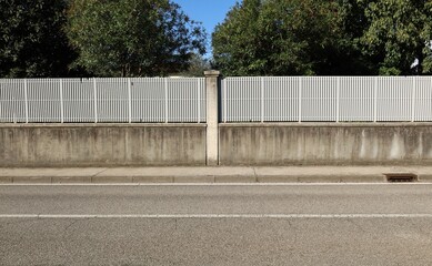 Wall Mural - Fence made of concrete wall with white railing on top. Decorative column in the middle. Trees on behind. Cement tiled sidewalk and street in front. Background for copy space.