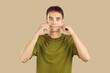 Close up studio portrait of young grimacing boy student wearing casual green t-shirt having fun pulling back his cheeks isolated on a beige background. People emotions, facial expression concept.