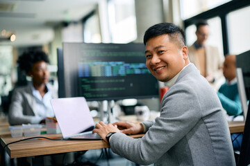 Happy Asian businessman working on laptop in office and looking at camera.